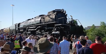 Una locomotora de vapor negra con la inscripción X4014 y una pequeña bandera estadounidense avanza sobre vías en un entorno de árboles verdes