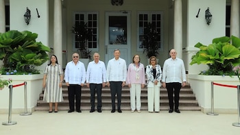 Siete funcionarios, tres mujeres y cuatro hombres, posan de pie frente a un edificio blanco con columnas, escaleras y grandes ventanales. La fachada tiene plantas a los lados