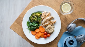 Un plato blanco con pollo, brócoli, espinacas, zanahorias, tomates y quinua. Junto al plato hay un vaso de agua con limón, auriculares y una banda elástica.
