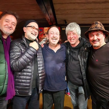 Cinco hombres abrazados y sonrientes en un recinto interior con techo de madera