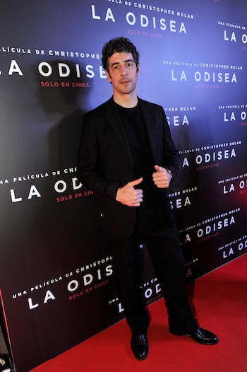 Un hombre vestido con traje negro y camiseta negra está de pie sobre una alfombra roja frente a un fondo negro con texto blanco y rojo