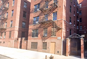 Edificio alto de ladrillo con múltiples ventanas, unidades de aire acondicionado, escaleras de emergencia y una entrada con rejas frente a una calle