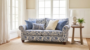Un sofá de dos plazas con estampado azul floral y cojines azules y crema. Una mesa auxiliar de madera con una lámpara blanca y una planta de helecho. Ventanas con cortinas.