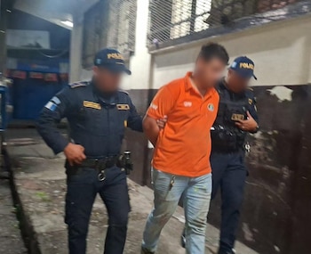 Dos agentes de policía con uniforme azul conducen a un hombre con polo naranja y rostro borroso