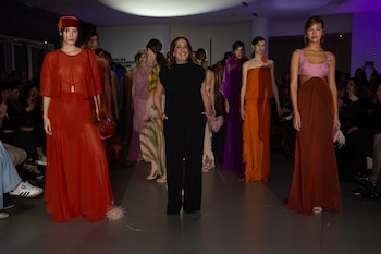 Modelos sobre una pasarela con vestidos de tonos rojos, naranjas y púrpuras junto a una mujer vestida de negro en una sala iluminada