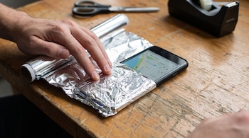 Manos de una persona que envuelve un teléfono celular con papel de aluminio sobre una mesa de madera con tijeras y cinta adhesiva.