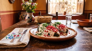 Dos chiles poblanos con crema blanca, granada y perejil en un plato de barro sobre una mesa de madera junto a cubiertos y flores.