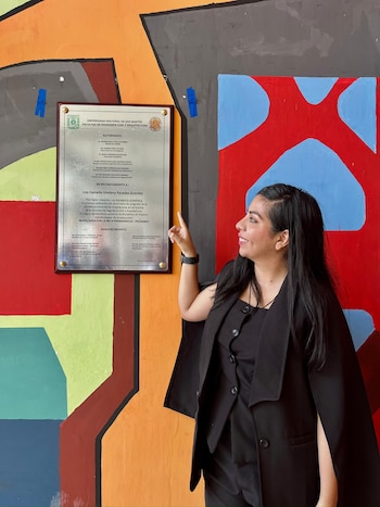 Una mujer de cabello oscuro y vestimenta negra sonríe y señala una placa de reconocimiento con texto en una pared con murales abstractos de colores