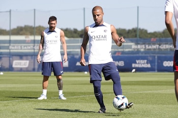 Rafinha Alcântara chuta com o pé direito no campo verde, com Lionel Messi atrás dele, ambos com uniforme de treino.