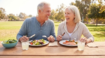 Una pareja sonriente de mediana edad sentada en una mesa de picnic en un parque. El hombre come una ensalada y la mujer salmón, con vasos de limonada y servilletas.