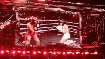 Duas boxeadoras com capacetes e luvas frente a frente em um ringue com cordas brancas, iluminado por luzes vermelhas e uma tela digital.