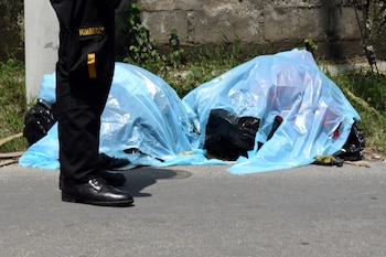 Piernas de un bombero con uniforme negro junto a dos formas cubiertas con plásticos azules y negros sobre el asfalto