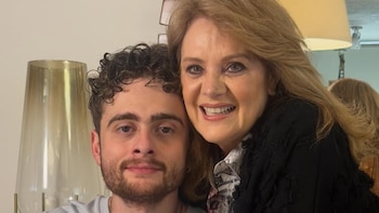 Primer plano de Erika Buenfil, una mujer rubia de mediana edad sonriendo, abrazando a un joven con cabello rizado oscuro y barba. Ambos miran a cámara