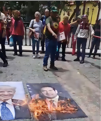 Una multitud de personas en una plaza pública observa dos retratos en el suelo, uno de Joe Biden, que están siendo consumidos por el fuego