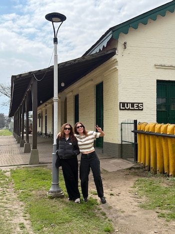 Dos mujeres con gafas de sol posan junto a un farol frente a un edificio antiguo de ladrillos con un cartel que dice Lules