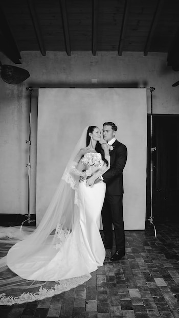 Fotografía en blanco y negro de una pareja vestida de boda, ella con velo y ramo, él con traje, abrazados frente a un fondo liso