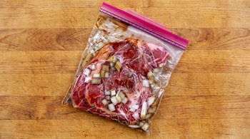Bolsa de plástico con un corte de carne cruda, cebolla picada y hierbas sobre una tabla de madera de cocina.