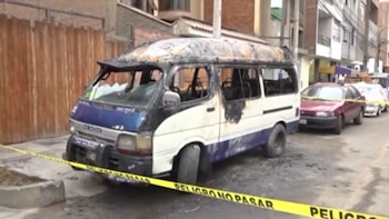Combi de transporte público totalmente calcinada en una calle urbana, rodeada por cinta de seguridad amarilla con la frase 'PELIGRO NO PASAR'