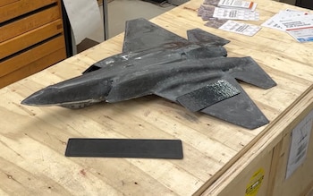 Maqueta de avión de combate en color oscuro sobre una superficie de madera, con una pieza rectangular negra y varios folletos impresos