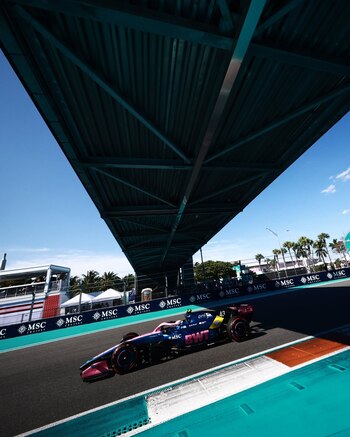 Un auto de Formula 1 rosa y azul con el número 43 en la pista bajo un puente, con cielo azul claro y palmeras al fondo durante el GP de Miami