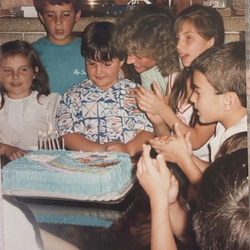 Grupo de niños y un adulto sonrientes alrededor de un pastel de cumpleaños azul con velas encendidas, en una foto de archivo vintage