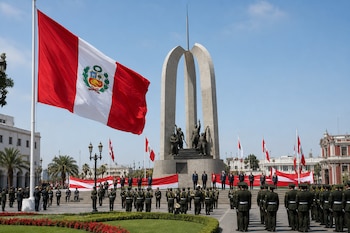 Una gran bandera de Perú flamea junto al monumento conmemorativo en una plaza pública donde autoridades y militares están reunidos.