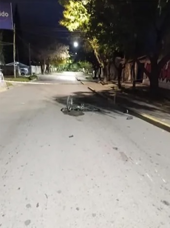 Vista nocturna de una calle oscura con una bicicleta tirada en el asfalto y manchas oscuras; árboles y una cinta amarilla en el fondo bajo la luz de un farol