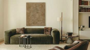 Vista frontal de una sala de estar moderna con un sofá de terciopelo verde oliva, cojines peludos, mesas redondas, un cuadro abstracto y parte de un sillón de cuero marrón.