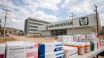 Vista exterior del edificio IMSS-Bienestar en una zona semi-rural, con cajas y frascos de medicamentos apilados en primer plano.