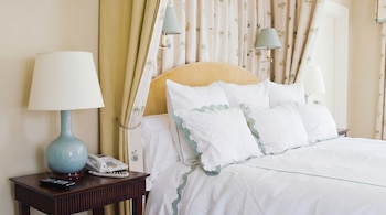 Cama con cabecero amarillo, ropa blanca, cojines borde azul, cortinas dosel estampadas, mesita de noche con lámpara, teléfono, control remoto.