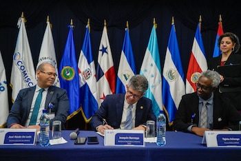 Tres hombres sentados en mesa. El del centro, Joaquin Barnoya, firma un documento. Banderas de países centroamericanos y COMISCA al fondo. Botellas de agua y micrófonos