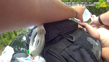 Vista en primera persona de un brazo vertiendo agua sobre el brazo y cuerpo de una niña, junto a una bolsa táctica negra, árboles y un vehículo