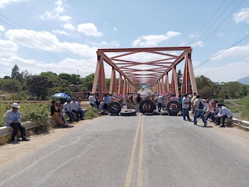Manifestantes bloquean un puente de celosía naranja con neumáticos y pancartas de "PARO AGRARIO". El cielo está despejado y hay vegetación a los lados del puente