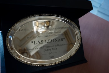 Una placa conmemorativa de plata ovalada con un borde dorado y un sello dorado superior, exhibida dentro de una caja negra. La placa tiene texto grabado