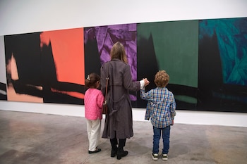 Una mujer y dos niños, de espaldas, observan cinco grandes paneles de pintura abstracta con colores como naranja, morado y verde en una galería