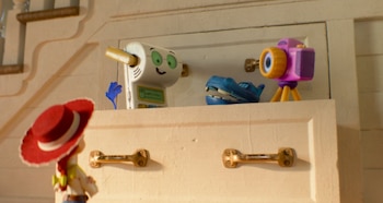 La figura de Jessie, un robot blanco con el nombre Smarty Pants, un juguete de hipopótamo azul y una cámara púrpura sobre un mueble de madera claro