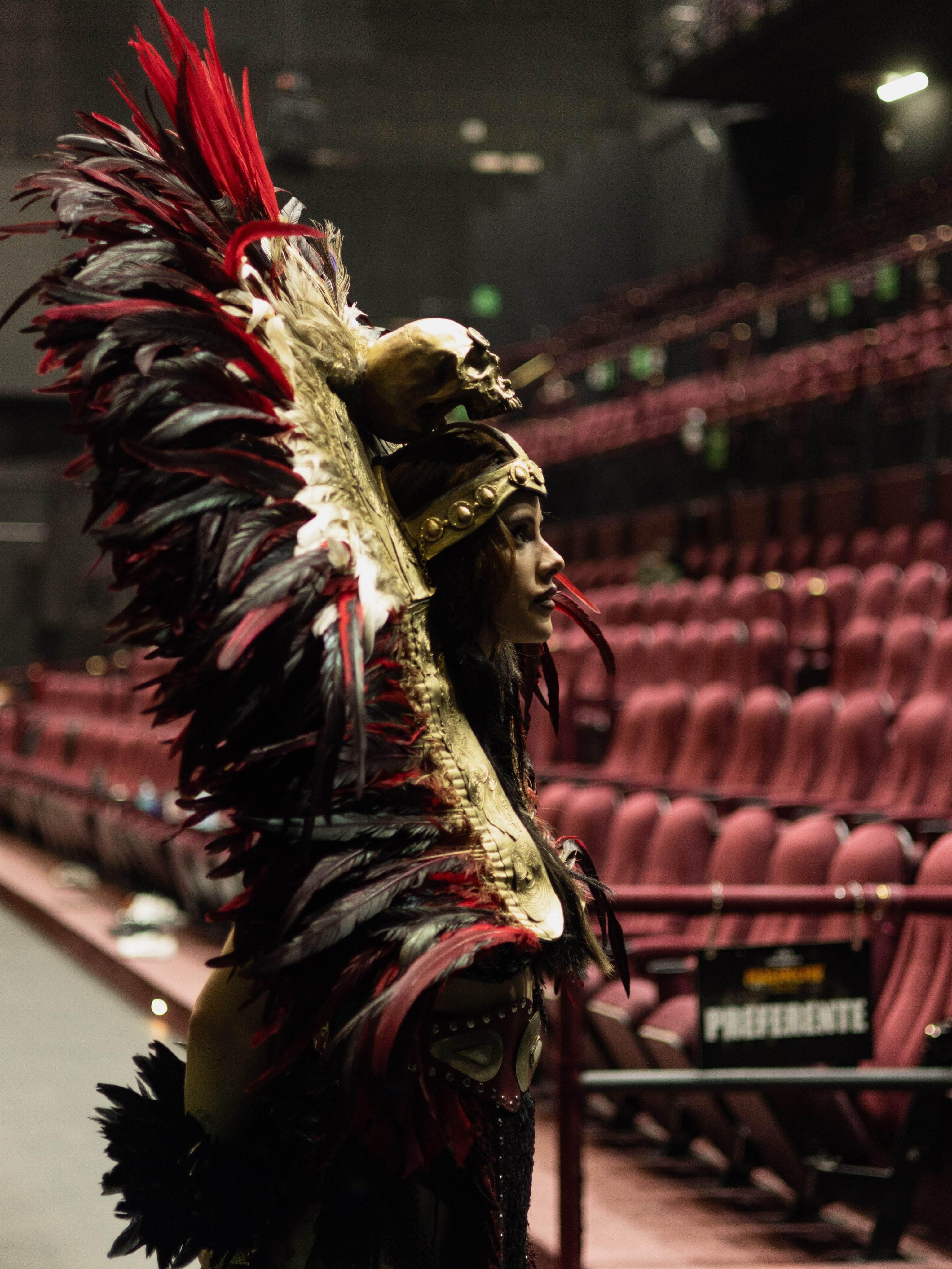 La actriz Gala Montes posa de perfil con un atuendo ceremonial de plumas y un adorno de calavera en el área de butacas de un teatro. (@malinchethemusical/Instagram)