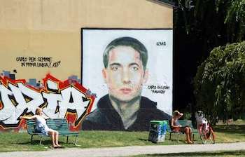 Un mural del rostro de un hombre joven con texto en italiano sobre una pared clara. Dos mujeres sentadas en bancos, un cubo de basura, una bicicleta roja y árboles