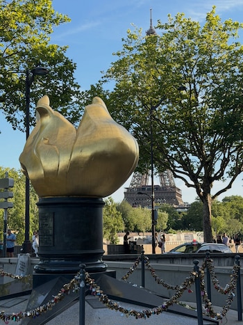 Escultura dorada de la Llama de la Libertad sobre pedestal. Torre Eiffel de fondo entre árboles. Cielo azul, peatones, un automóvil y vallas con candados