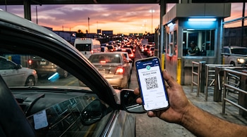 Una mano sostiene un teléfono con un código QR en la pantalla frente a una fila de automóviles y una caseta de control al atardecer.