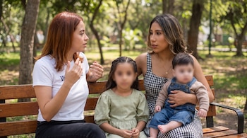 Dos mujeres y dos niños sentados en un banco de madera en un parque; los rostros de los menores están pixelados.