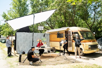 Un equipo de filmación graba en un set al aire libre con una mujer en una mesa, rodeada de luces, un micrófono boom y un RV amarillo vintage al fondo