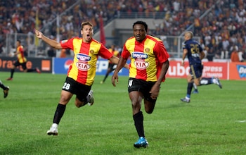 Dos futbolistas en camisetas rojas y amarillas con números 28 y 14 corren sobre césped verde en un estadio, con espectadores y vallas publicitarias al fondo