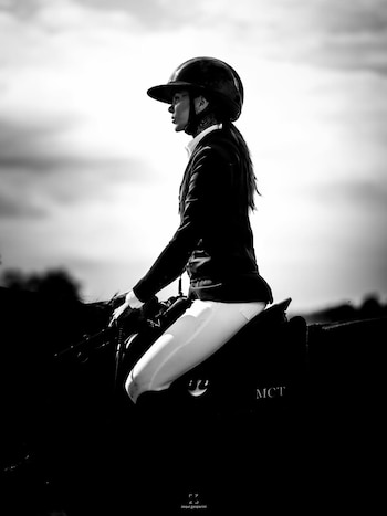 Fotografía en blanco y negro de Cande Tinelli a caballo, de perfil, vistiendo casco y traje de equitación. El cielo nublado se ve al fondo