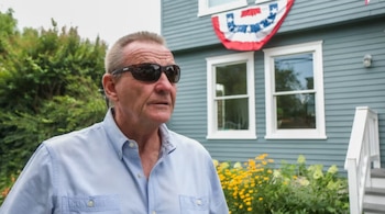 Primer plano de un hombre maduro con gafas de sol oscuras y camisa azul claro, de pie al aire libre frente a una casa de color gris azulado con una bandera estadounidense decorativa