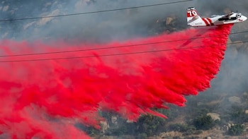 Avión blanco y rojo con el número 70 en la cola descarga retardante de fuego rojo brillante sobre un paisaje humeante con vegetación y líneas eléctricas