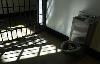 Una celda de prisión muestra rejas de metal a la izquierda, proyectando sombras en el suelo. A la derecha, un inodoro y un lavabo de acero inoxidable