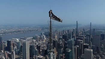 Vista aérea de dos personas en la cúspide de una torre alta, desplegando una pancarta negra, sobre un panorama de ciudad con rascacielos y un río