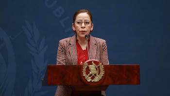 Mujer con anteojos y chaqueta a cuadros habla ante un micrófono en un podio de madera con el escudo de Guatemala