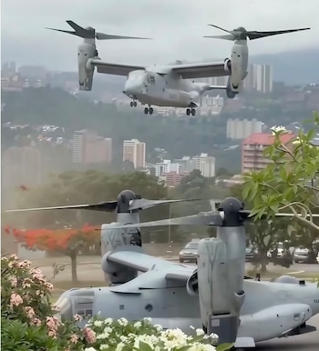 Dos aviones militares de ala basculante, uno volando sobre edificios urbanos y otro parcialmente visible en tierra detrás de arbustos con flores